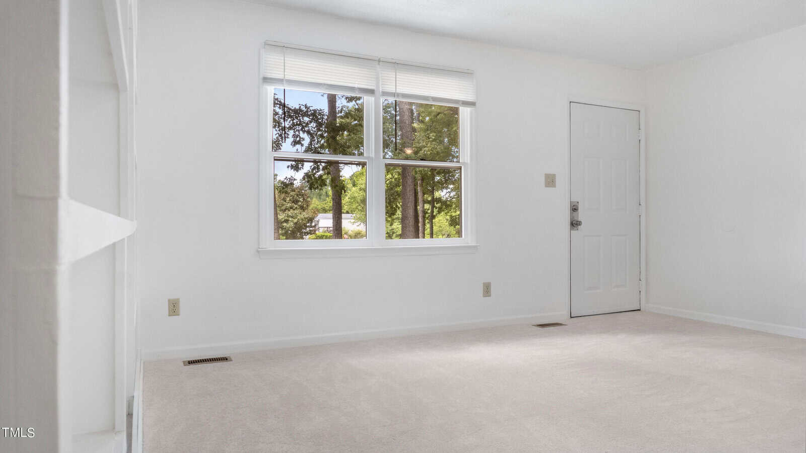 A bright, empty room with white walls and a light carpeted floor. The room has a window showing trees outside and a white door with a silver doorknob. Natural light illuminates the space.