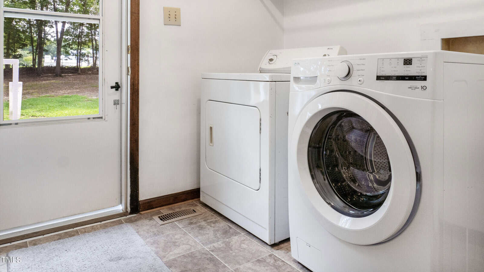 A laundry room with a white front-loading washing machine and a white top-loading dryer side by side. The room has white walls, a windowed door leading outside, and a tiled floor. A floor vent is visible next to the machines.