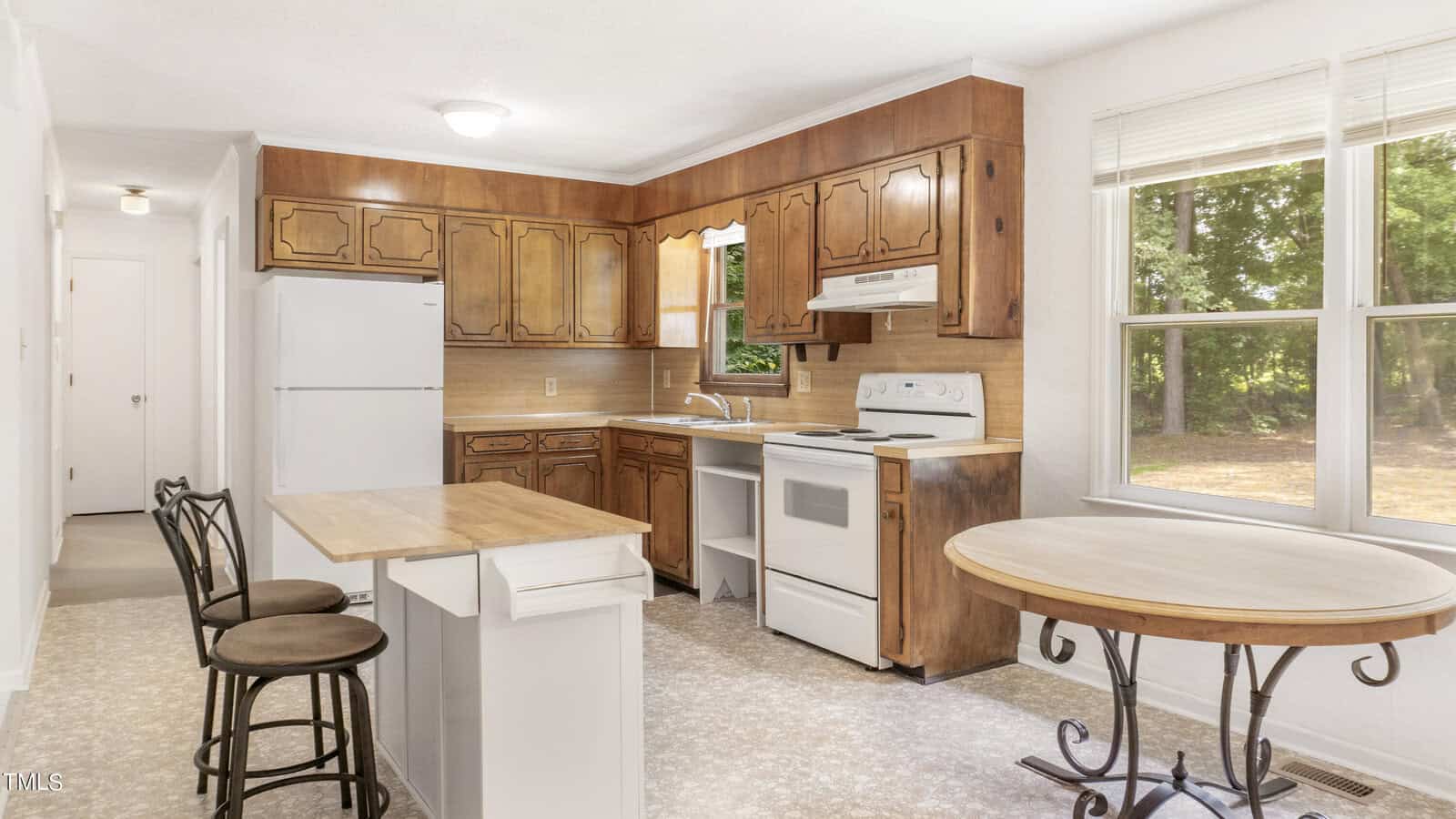 A kitchen with wooden cabinets and white appliances, including a refrigerator and stove. A small island with two bar stools is in the foreground, along with a round table. Two windows on the right side let in natural light.