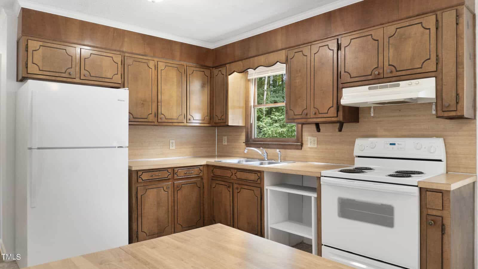 A kitchen with wood cabinets and a white refrigerator. It features an electric stove and oven, a stainless steel sink, and a window above the sink. The countertop and walls are a light, neutral color. A small section is open for additional storage.
