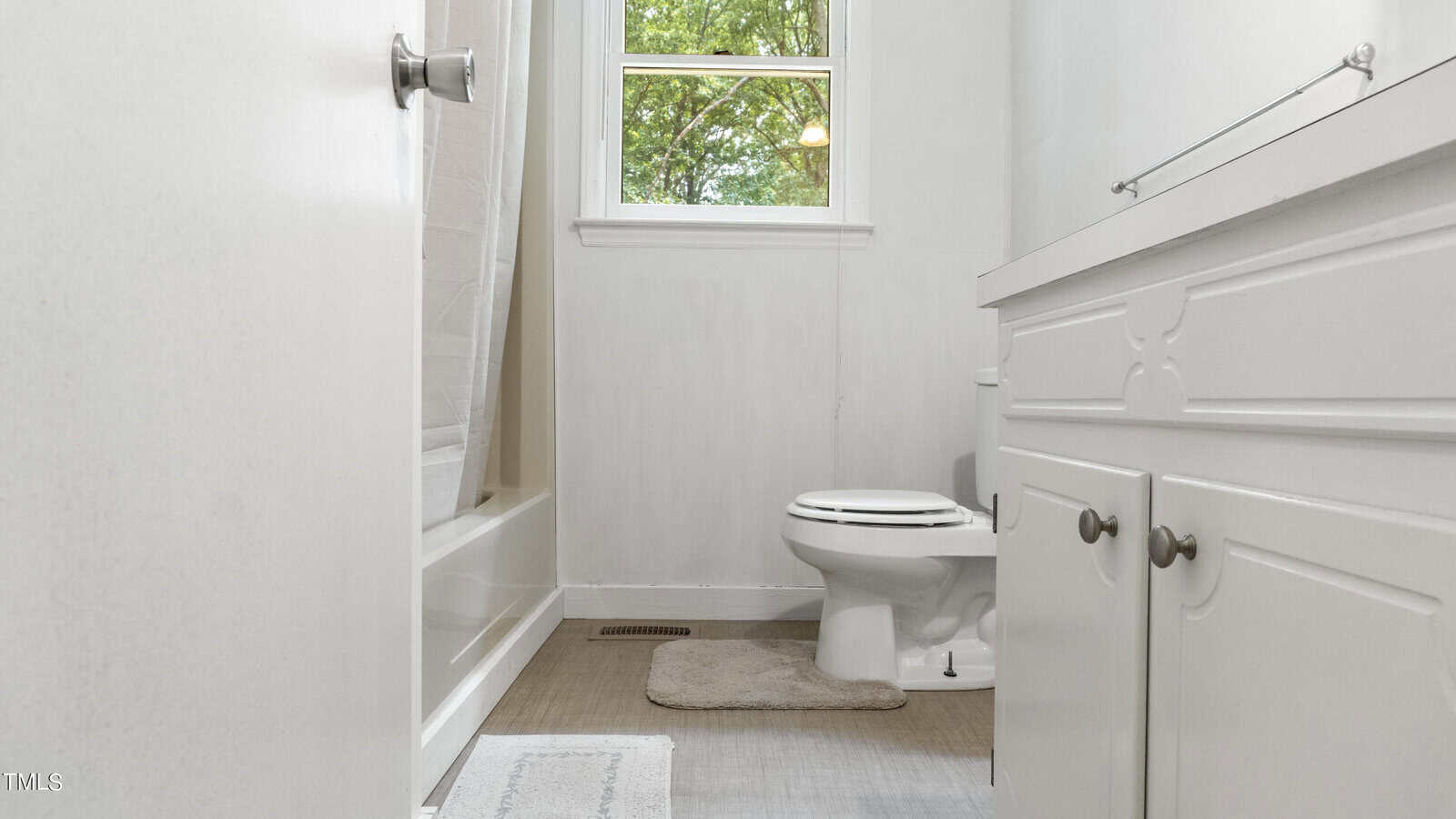 A small bathroom with white walls features a bathtub with a curtain, a toilet, and a sink with a cabinet. There is a window above the toilet, letting in natural light. The floor is covered in light-colored tiles, and a rug is placed near the tub.