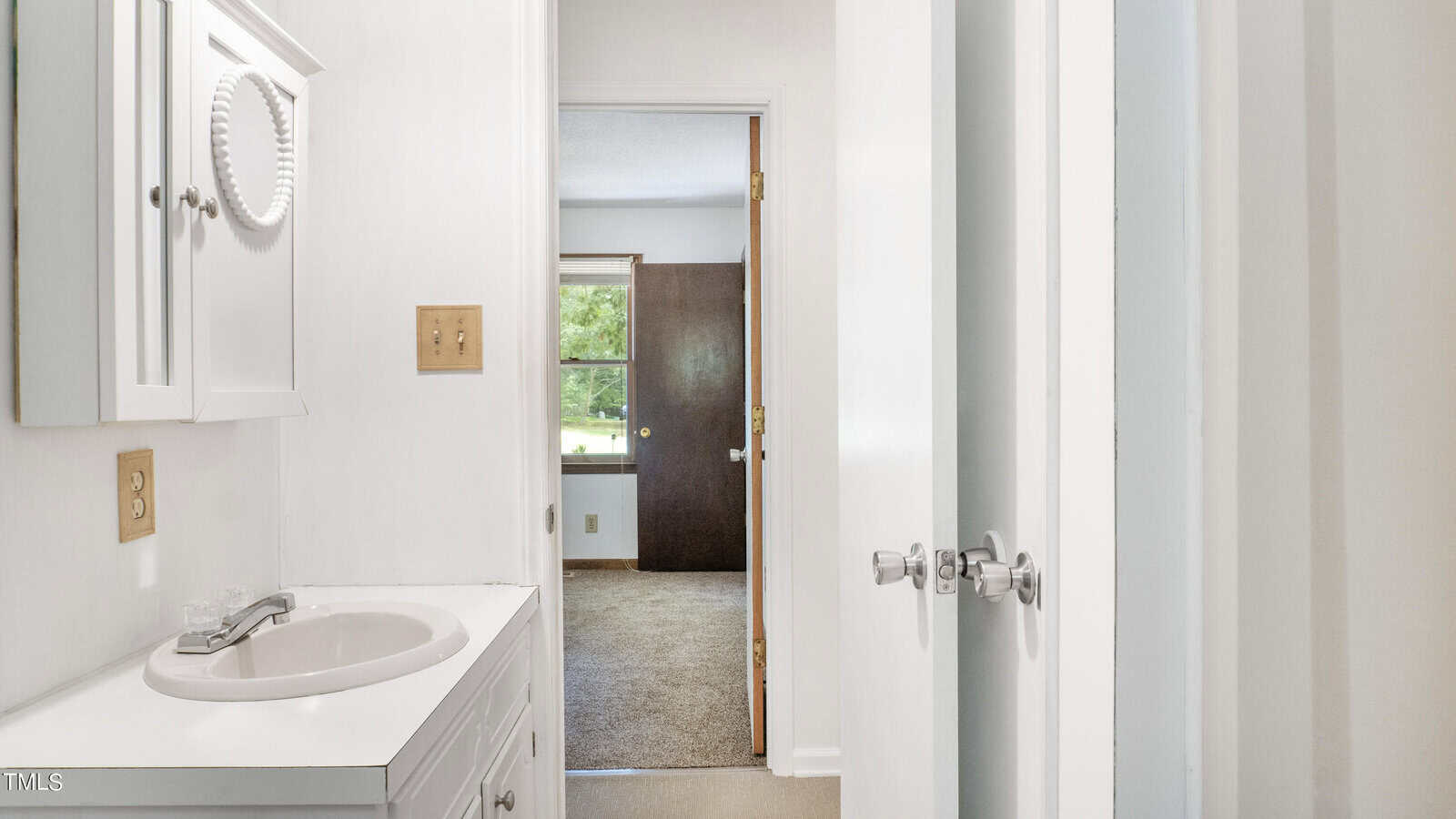 A narrow, white bathroom with a small sink and mirrored cabinet on the left. The space leads to a door opening into a carpeted room with a window facing a green outdoor view. Two silver knobs are visible on a closed door on the right.