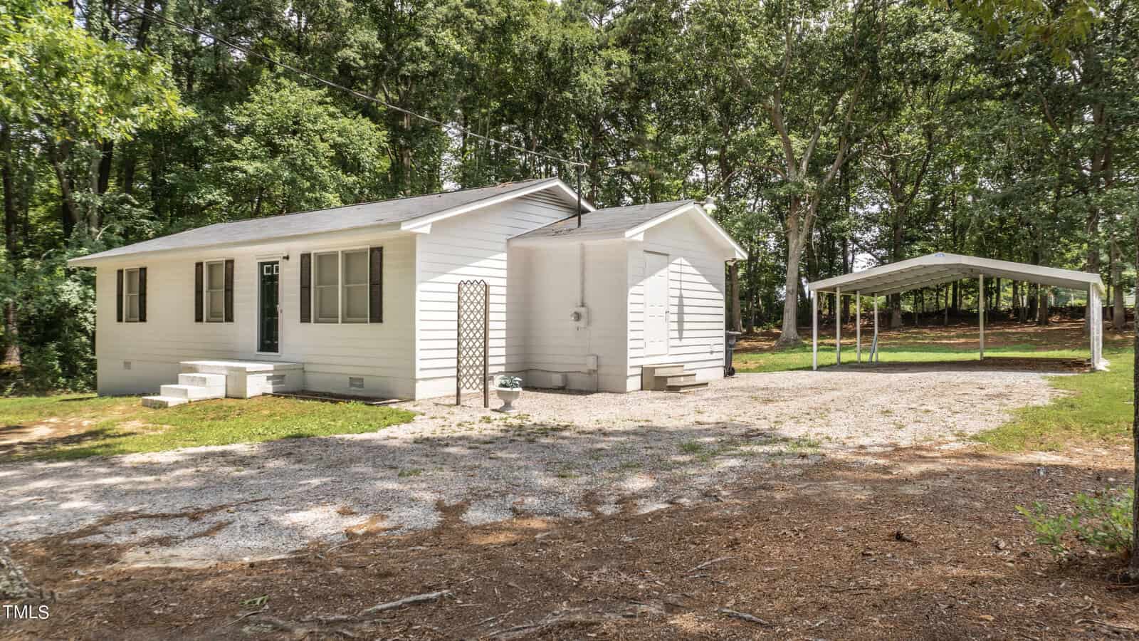 A small white house with a green door is situated among trees. It has a gravel driveway leading to a detached carport on the right. The surrounding area is wooded, providing shade and greenery.