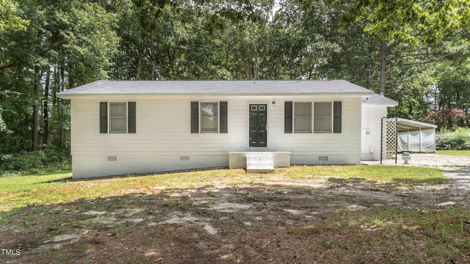 Nestled in a clearing of trees on 113 S Lake Lodge Road, Henderson lies a small, single-story white house. It features a dark green door and shutters, with a simple front stoop and gravel driveway. A carport sits to the right, offering shelter for your vehicle.