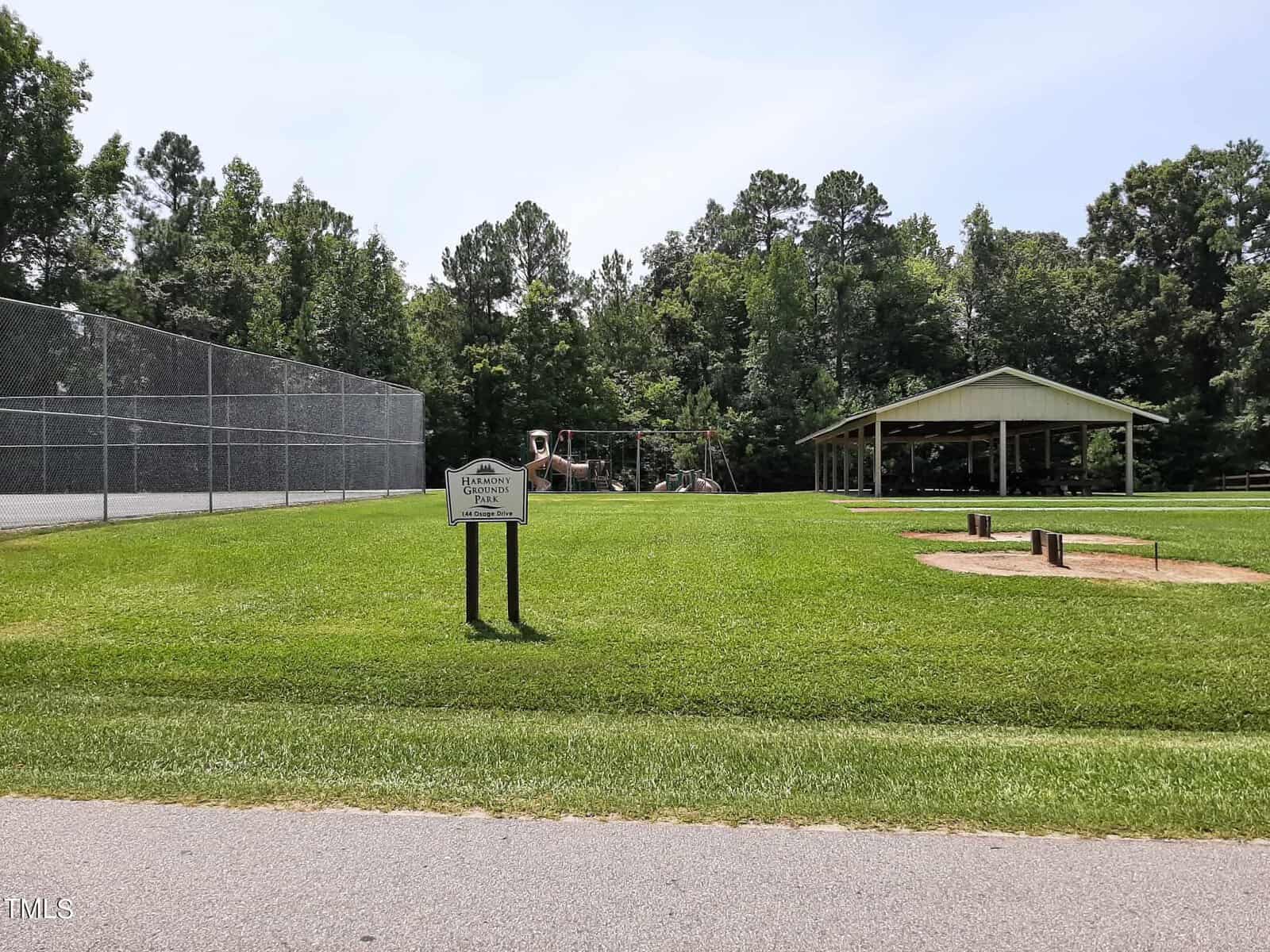 A grassy park area with a tennis court on the left, a playground in the center, and a covered picnic pavilion on the right. A wooden park sign is visible in the foreground. Trees line the background.