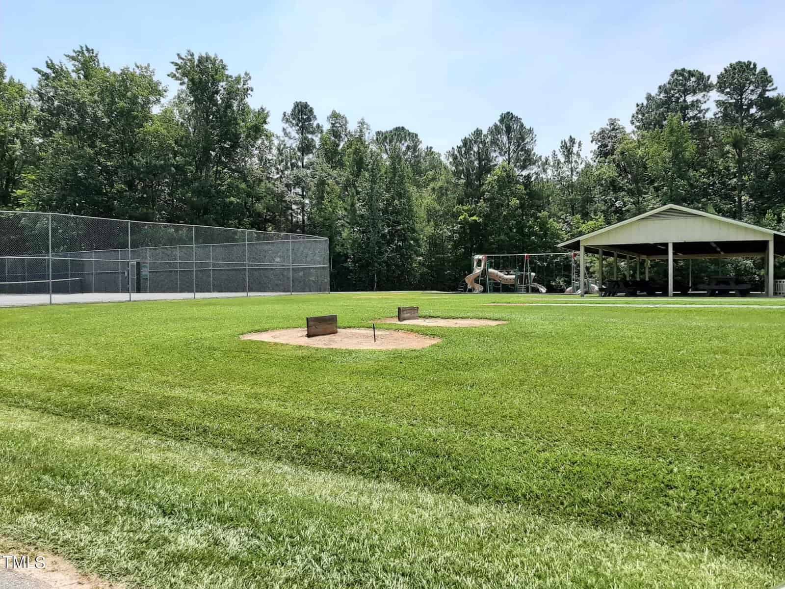A grassy park area with two horseshoe pits in the foreground, a chain-link fence and tennis courts to the left, and a covered pavilion with a playground in the background surrounded by trees.
