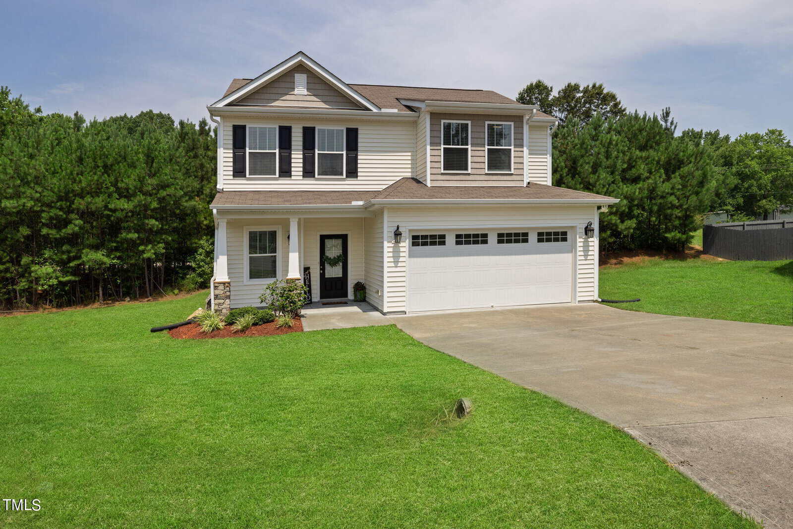 A two-story suburban house with beige siding and white trim, featuring a two-car garage and a well-maintained lawn. The house is bordered by a lush green hedge of trees, and the driveway curves gently to the right.