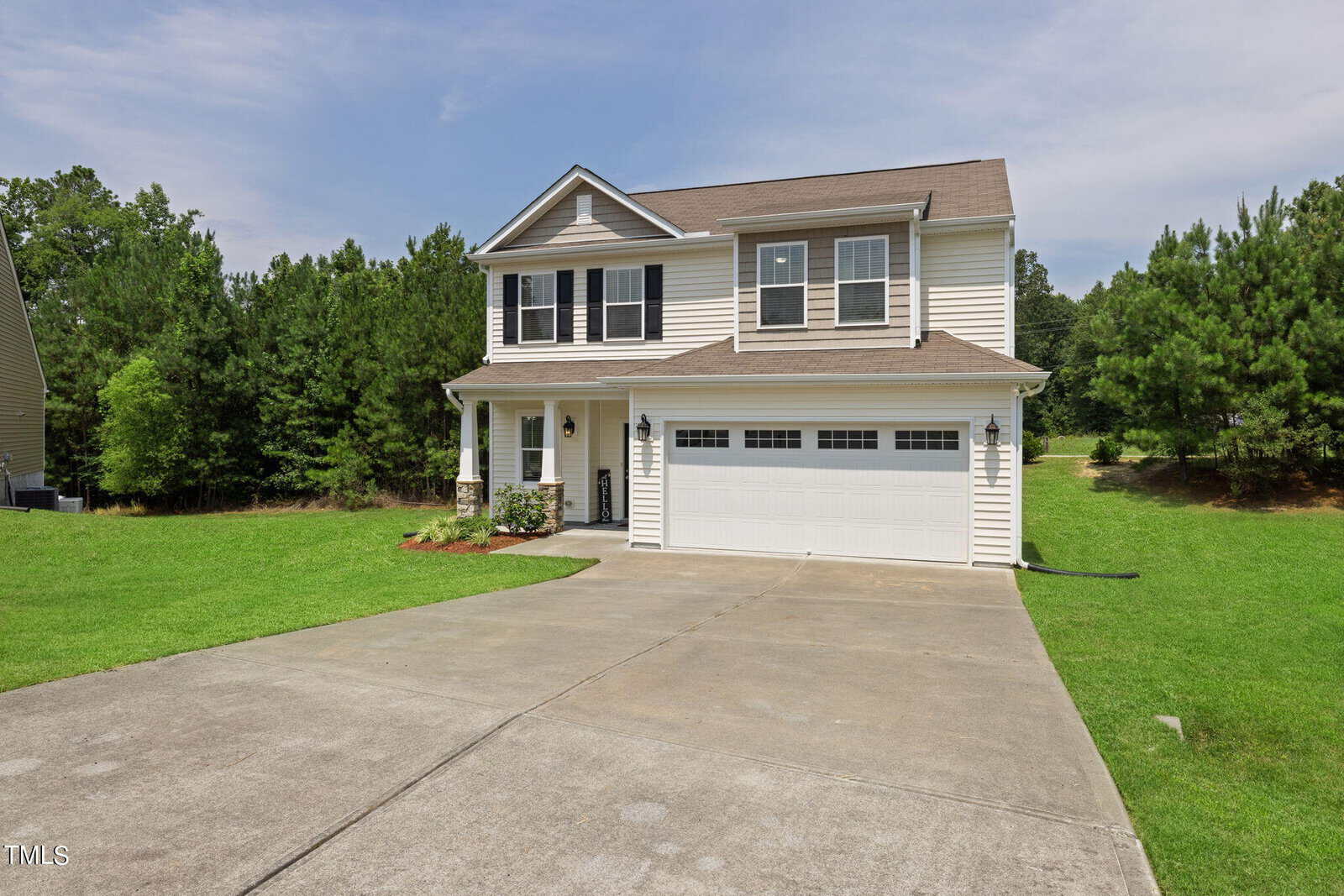 A two-story house with beige siding and dark shutters, featuring a double garage and a neatly maintained lawn. Surrounded by trees, the home sits under a blue sky with a concrete driveway leading up to it.