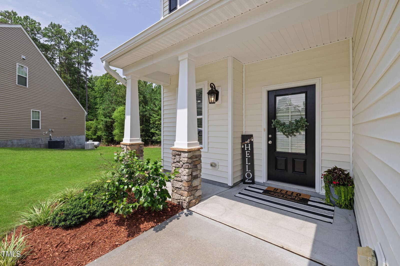 Front porch of a house with a "Hello" sign and a doormat reading "Hello." The black door features a wreath. White columns and stone accents support the porch. A plant is near the entrance, and there is a green lawn in the background.