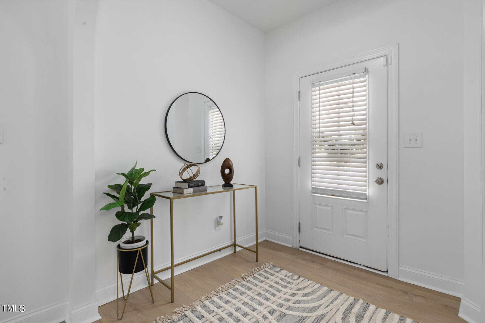 A minimalist entryway with a white door and blinds. A small table with a gold frame holds a potted plant, a round mirror, a decorative sculpture, and a book. A patterned rug covers the light wood floor.