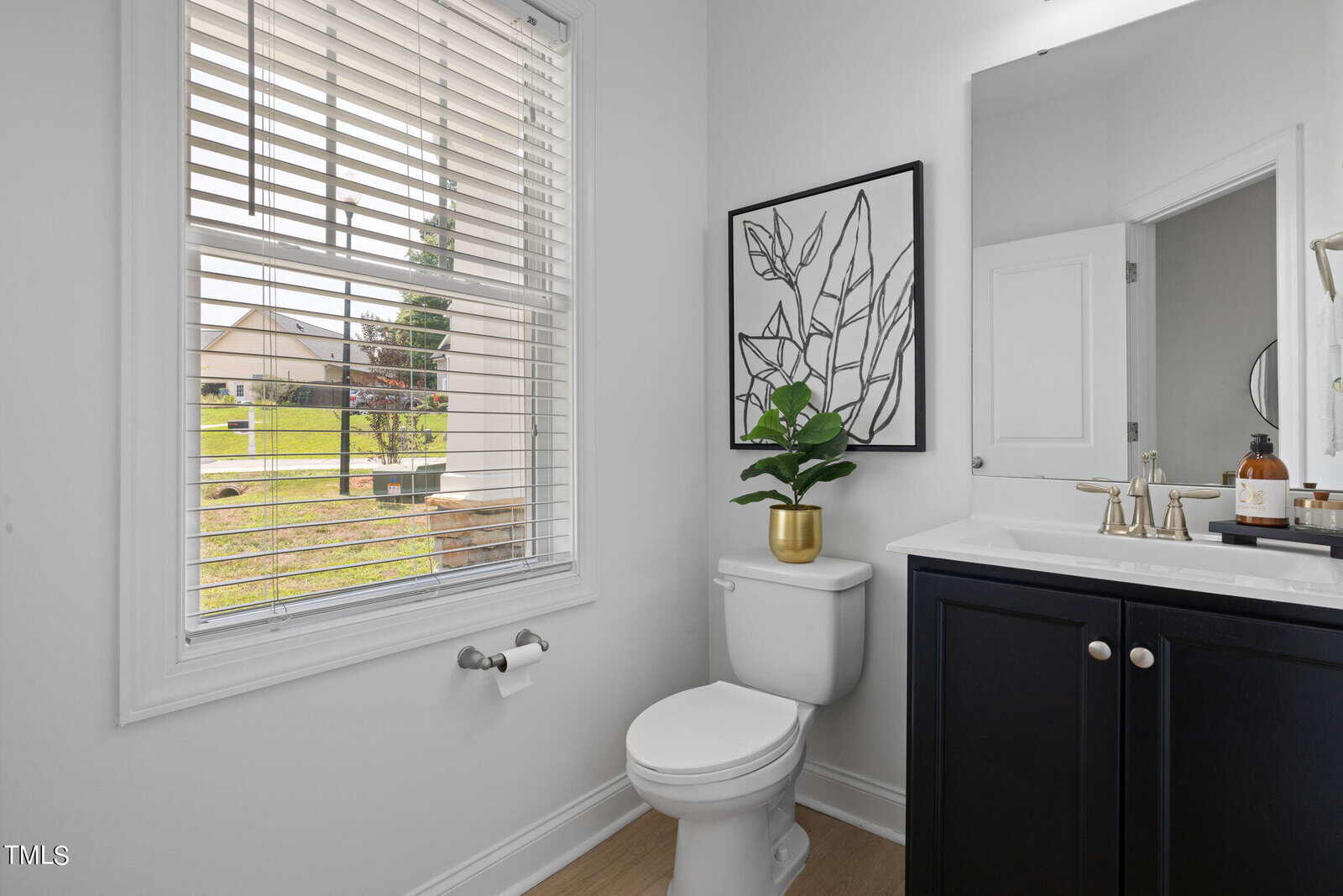 A modern bathroom with white walls and wood flooring features a toilet, a white sink atop a dark vanity, and a decorative plant in a pot. A large window with blinds brings in ample natural light. A framed leaf artwork hangs above the toilet.