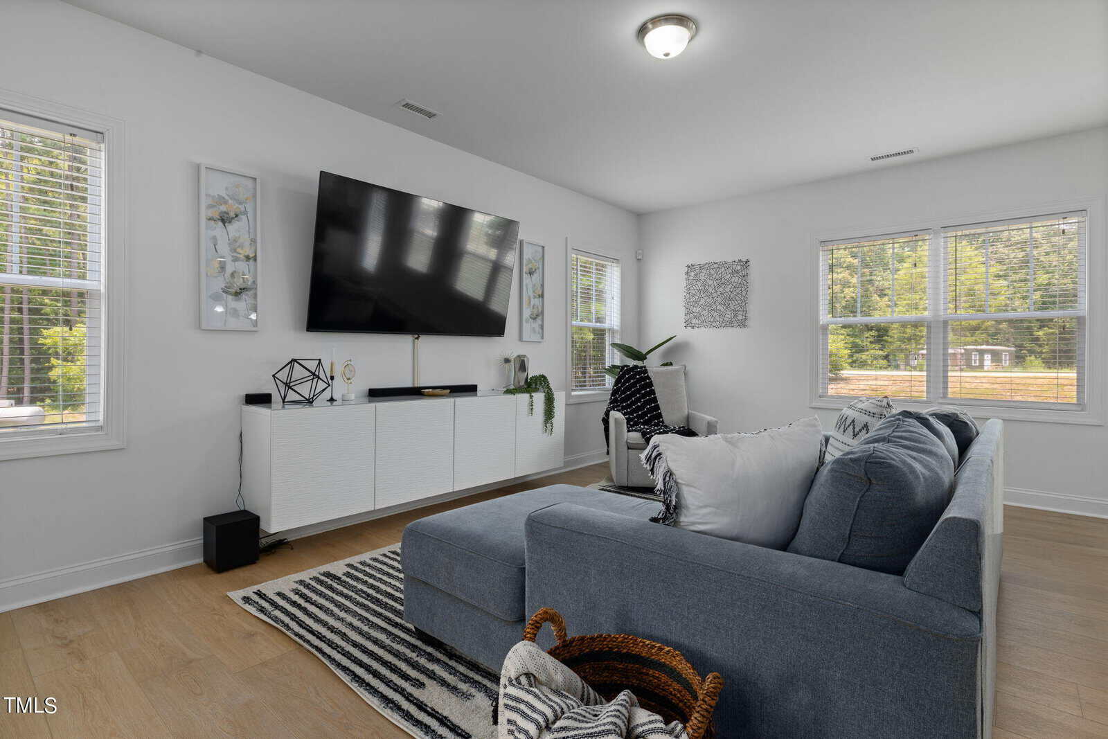 A modern living room with a large wall-mounted TV above a sleek white console. A gray sectional sofa faces the TV, adorned with cushions. Light floods the room through several windows, and various decorative items are neatly arranged.