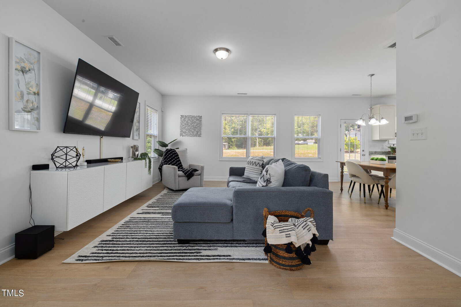 A modern living room with a large wall-mounted TV, white cabinets, and gray accent chairs. A gray sectional sofa is centered on a striped rug. Light pours in from windows, and a view of a dining area is visible in the background.