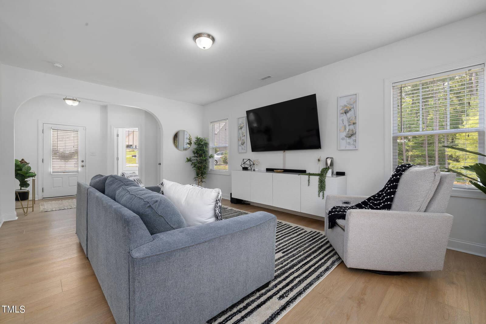 A modern living room with a gray sofa, a light armchair with a black and white throw, and a wall-mounted TV. The room features wood flooring, a striped rug, and large windows with blinds, allowing natural light to brighten the space.