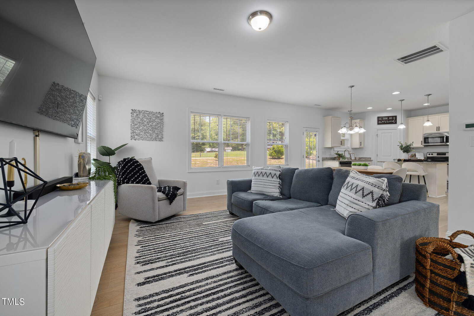 A modern living room with a gray sectional sofa, striped rug, and a wall-mounted TV. Large windows let in natural light. A dining area with a table and chairs is in the background, adjacent to a kitchen with white cabinets and stainless steel appliances.