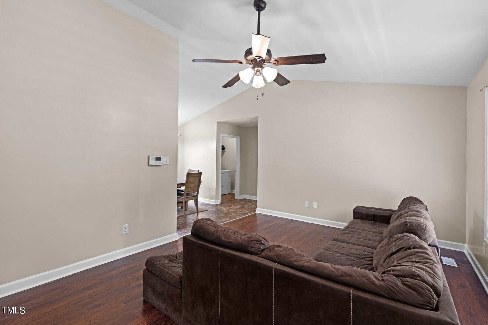 A living room with a brown sectional sofa on dark hardwood flooring. A ceiling fan with lights is above, and an open doorway leads to a dining area with a table and two chairs. Walls are painted in a light beige color.