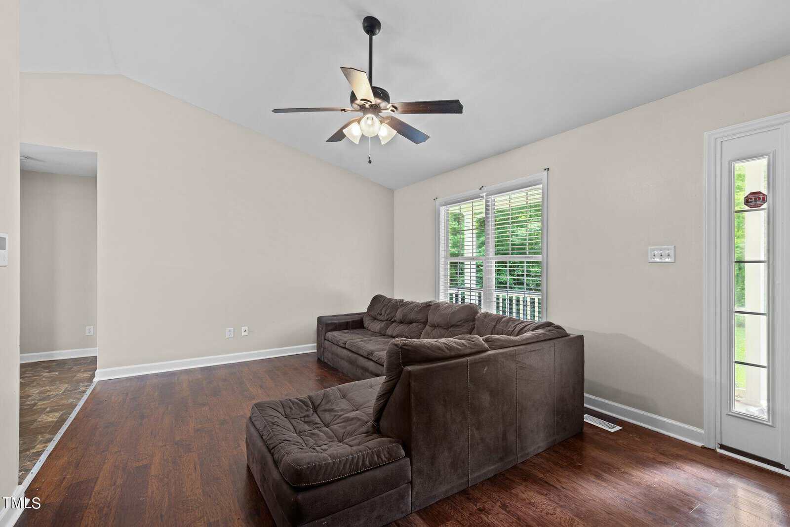 A living room with a brown sectional sofa on a hardwood floor. The room has beige walls, a ceiling fan with lights, and a large window with blinds that let in natural light. An open doorway leads to another area of the house.