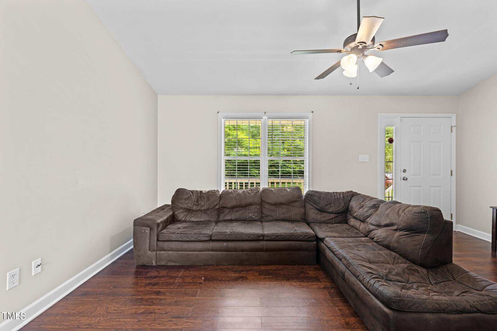 A living room with a large brown sectional sofa on dark wood flooring. The room has cream-colored walls, a ceiling fan with lights, two windows with blinds, and a door leading outside. Natural light streams in through the windows.