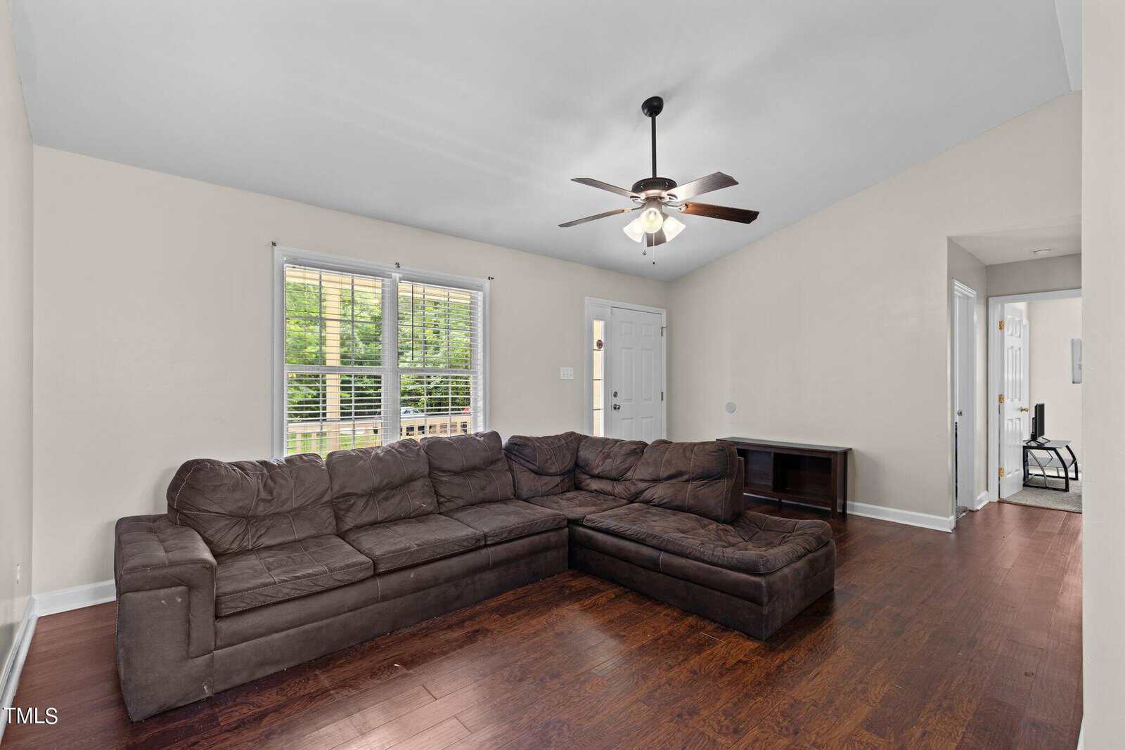 A living room with an L-shaped dark brown sofa on wooden flooring. A ceiling fan is overhead, and a large window with blinds lets in natural light. The walls are light-colored, and a small console table is against the far wall.