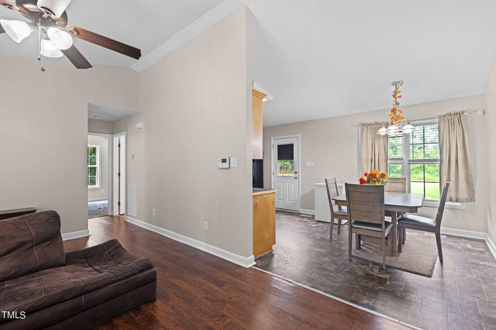 A modern living space features a ceiling fan and a large brown sectional sofa. Adjacent is a dining area with a wooden table, surrounded by chairs and topped with a fruit basket. Large windows allow natural light, and a door leads outside.