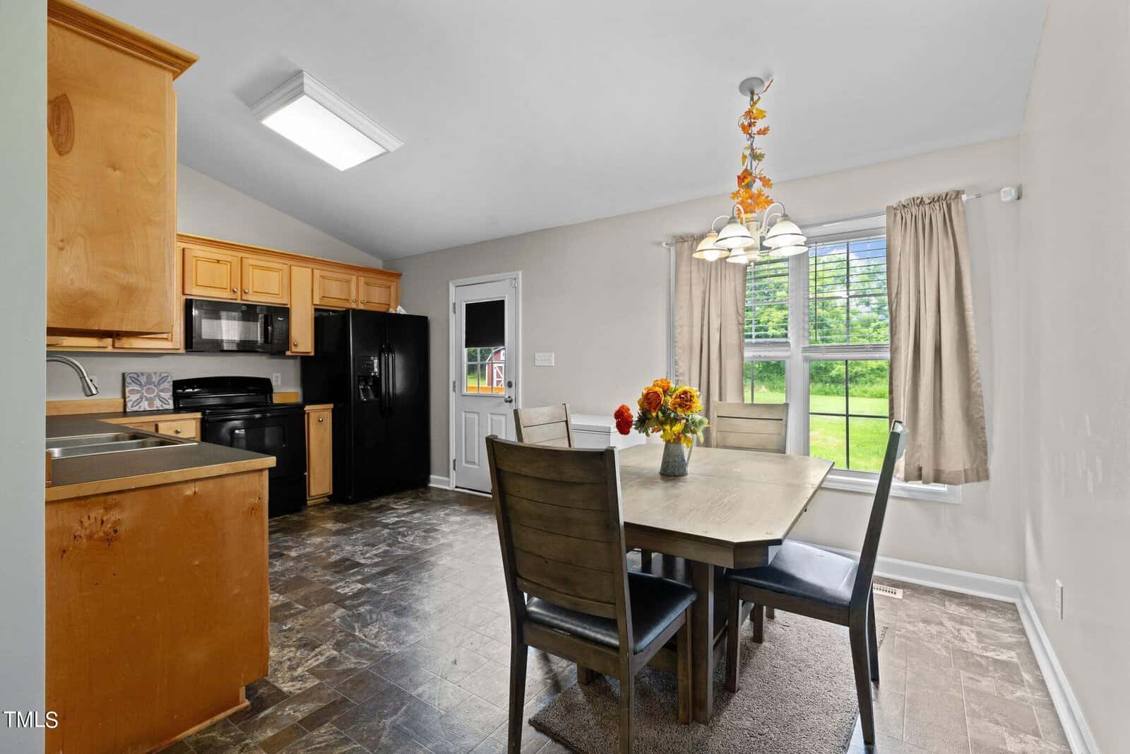 A bright kitchen and dining area with wooden cabinets, a black fridge and stove, and a dining table with four chairs. A vase of flowers sits on the table. There's a large window and a door leading outside, allowing natural light to fill the room.