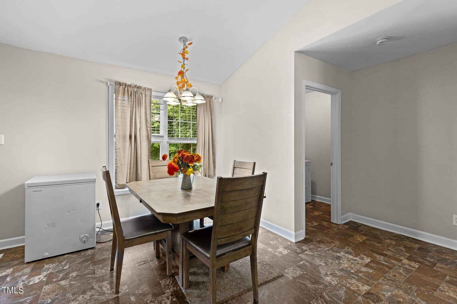 A dining room with a wooden table and four chairs, a floral centerpiece, and a light fixture above. The room has beige walls, a window with curtains, and a small white appliance against the wall. The floor is covered with textured tiles.