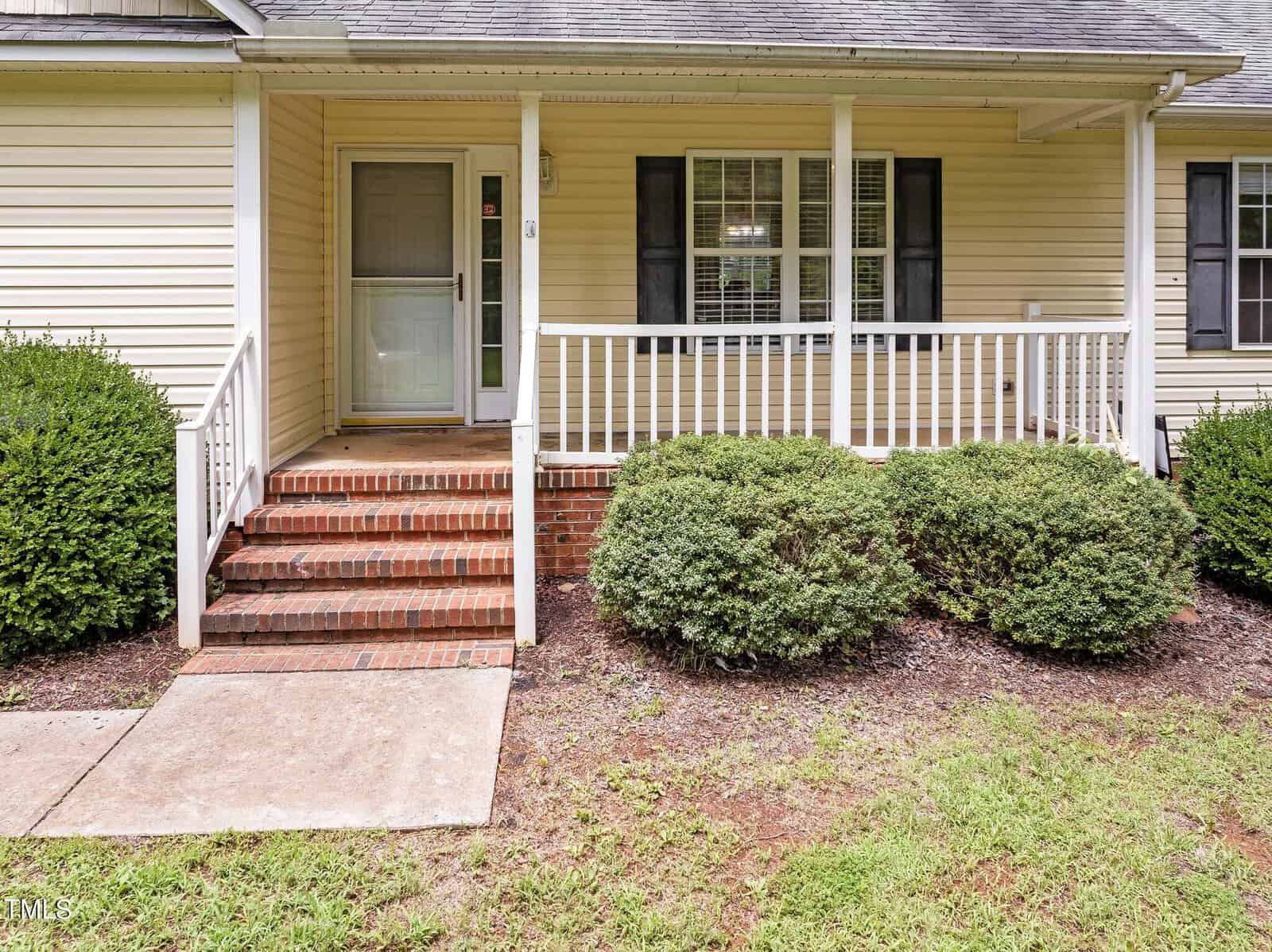 A cream-colored house with a small front porch and white railings. Three steps lead up to a front door. Two bushes are on either side of the steps, and a section of grass is visible in the foreground.