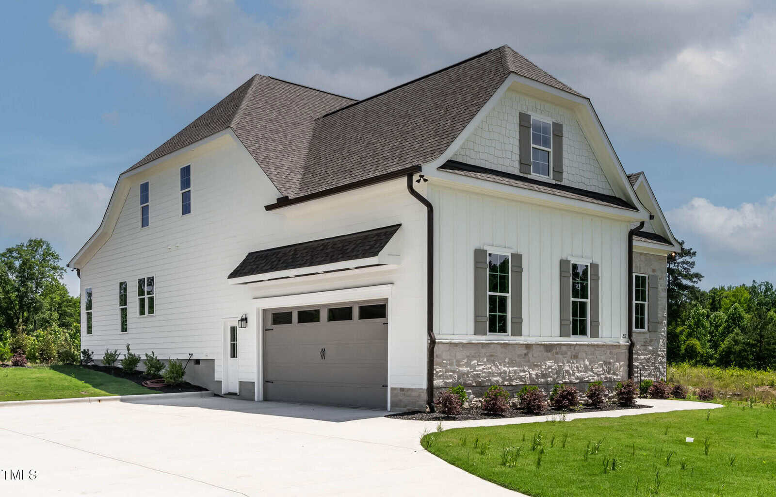 A modern two-story white house with gray roof shingles, a stone accent on the lower facade, large windows, and a double garage. The driveway and neatly landscaped yard are in front, with trees in the background.