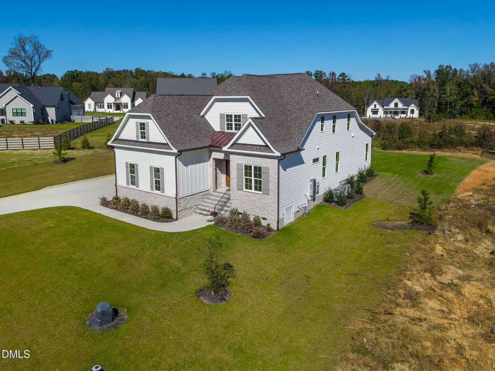 A large, two-story, light-colored house with a shingle roof sits on a spacious, well-manicured lawn with young trees. The home features multiple gables, a front porch with steps, and surrounding greenery.