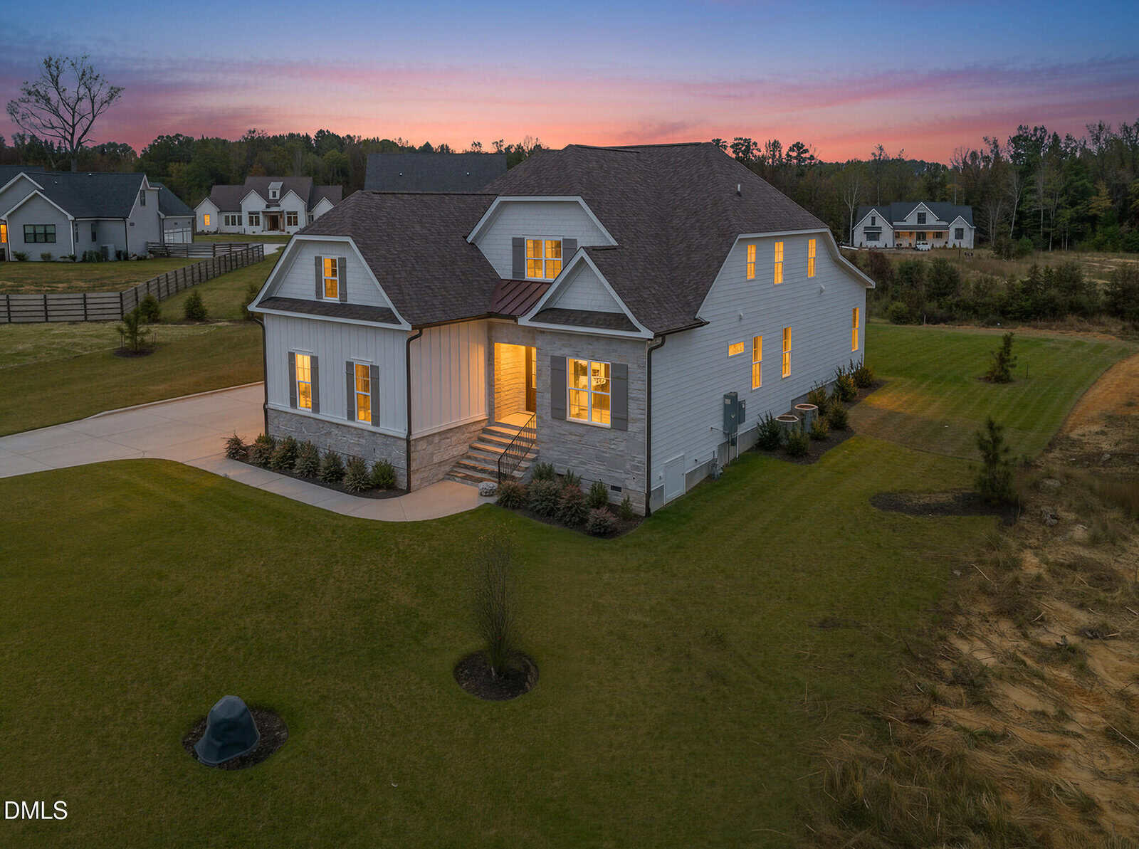 Two-story modern farmhouse with large windows lit from within, surrounded by a spacious lawn at sunset. Neighboring homes and trees are visible in the background under a colorful sky.