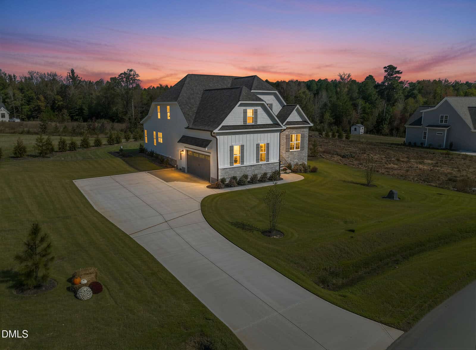 A modern two-story house with lights on, sits on a large, neatly mowed lawn with a long curved driveway at sunset. The sky glows pink and purple, and trees line the property in the background.