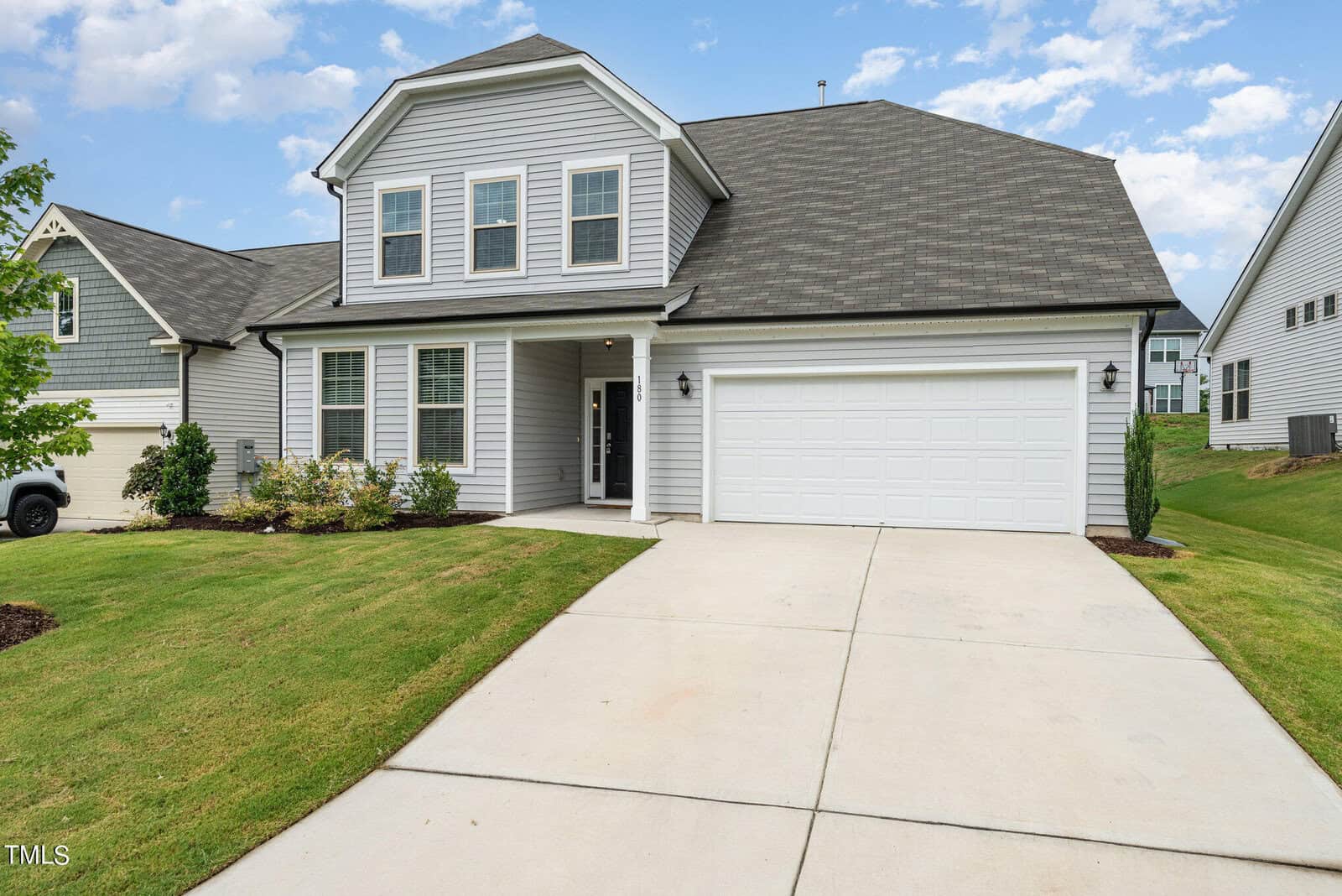 Two-story, light gray house with white trim, dark roof, double garage, and covered front porch. Neatly manicured lawn and shrubs line the driveway and front entrance on a clear day.