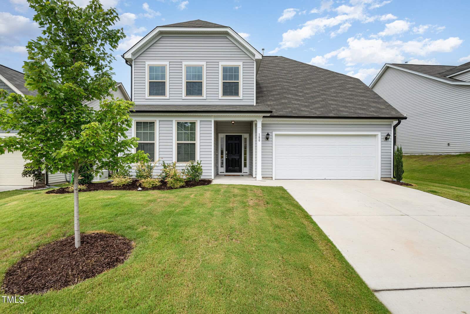 Two-story suburban house with gray siding, white trim, and black shutters, featuring a double garage, manicured lawn, small front tree, and a driveway under a partly cloudy sky.