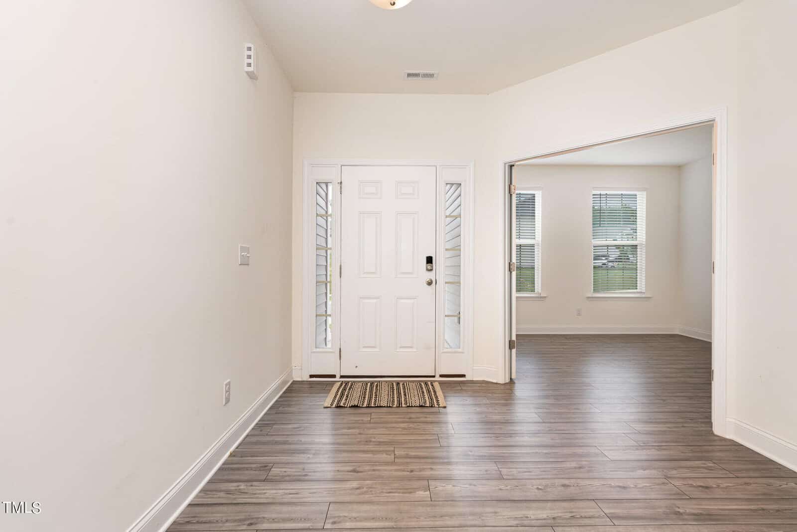 A bright entryway with light walls, wood-look flooring, a white front door with glass panels, and an adjacent room with large windows and similar flooring visible through double doors.