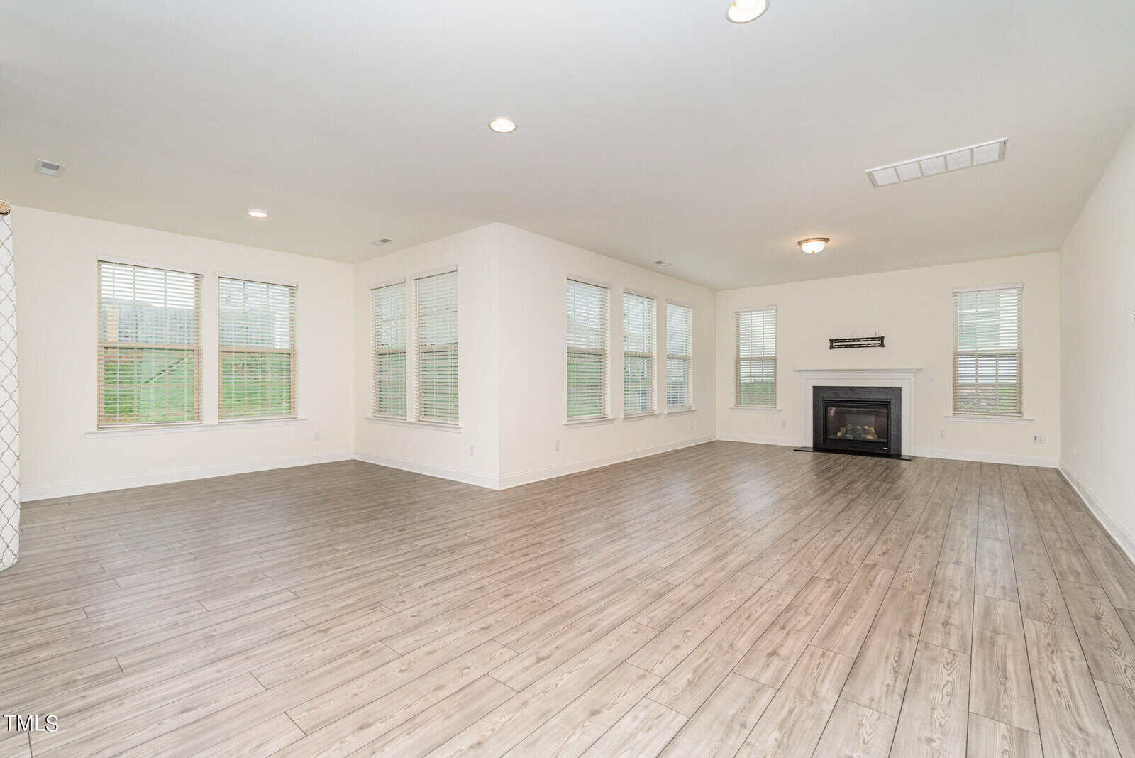 Spacious, empty living room with light wood floors, large windows letting in natural light, white walls, and a corner fireplace with a mantel. The room has recessed lighting and a bright, open feel.