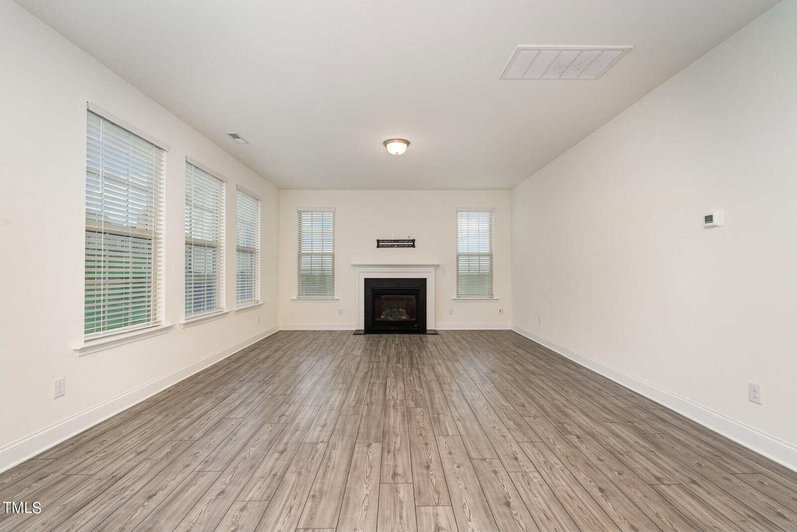 Spacious, empty living room with light-colored walls, wood-style flooring, large windows on two sides, and a fireplace centered on the far wall beneath a small wall-mounted sign. Ceiling light fixture is visible.