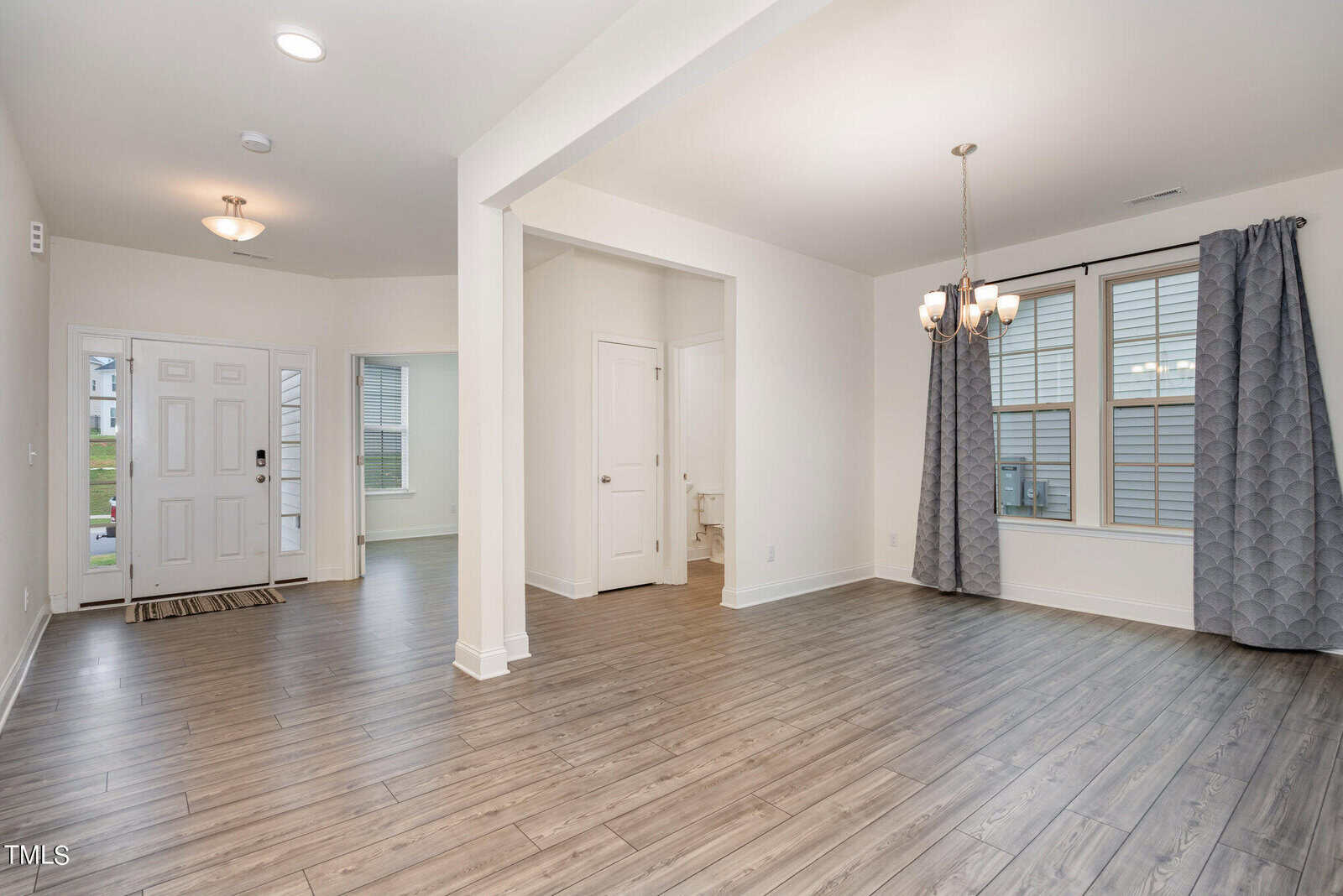 Open living area with wood flooring, light walls, a modern chandelier, and large windows with gray curtains. Entryway and hallway with white doors visible; natural light fills the space.
