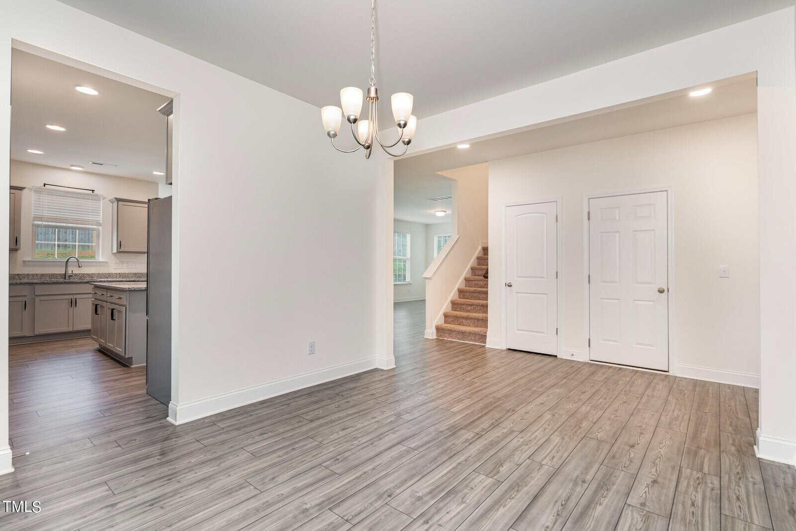 Open, modern home interior with wood flooring, white walls, a chandelier, two closed doors, visible staircase, and a kitchen area with gray cabinets and stainless steel appliances in the background.