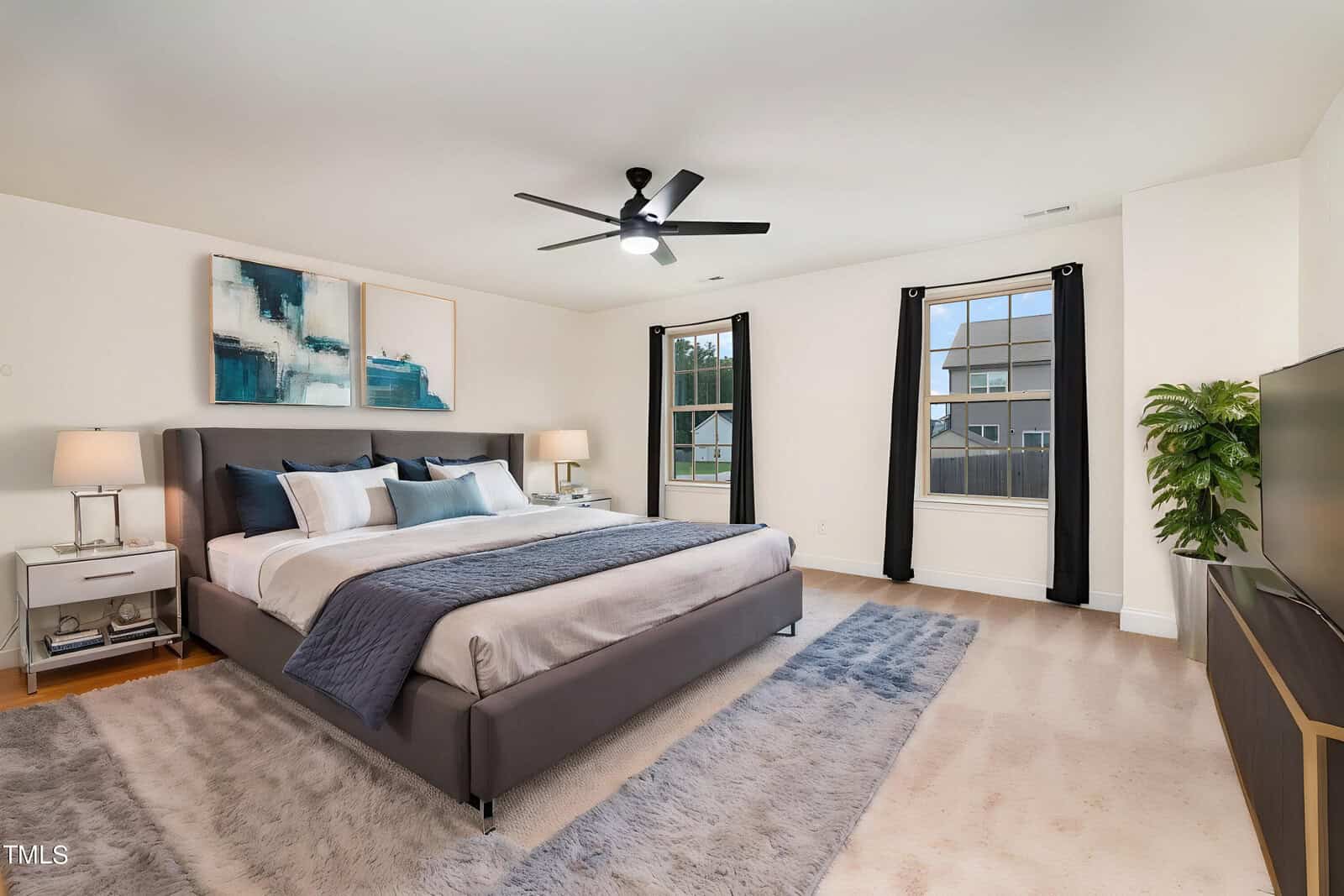 A spacious, modern bedroom with a large gray bed, white bedding, two nightstands with lamps, abstract wall art, a flat-screen TV, and two windows with black curtains letting in natural light.