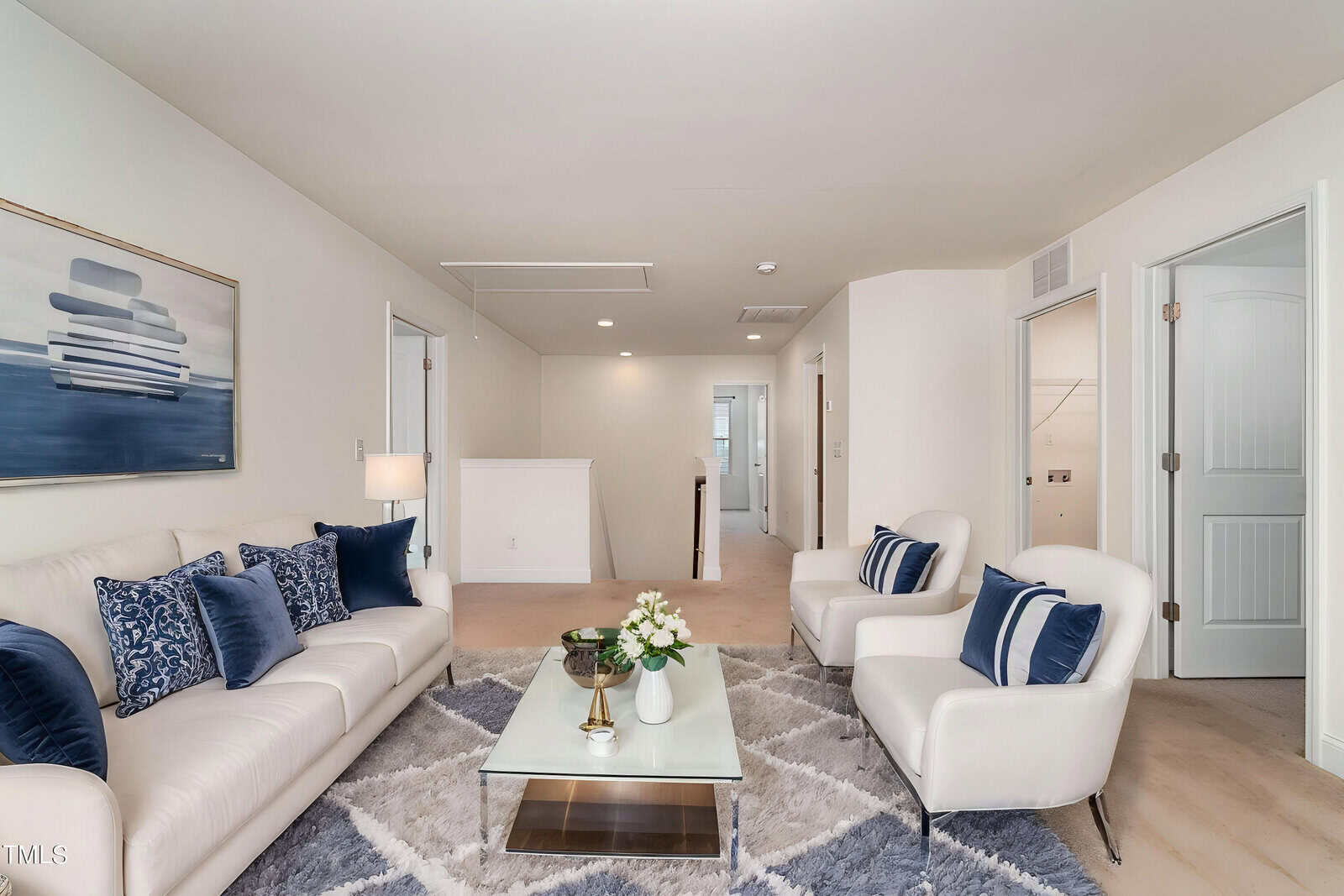 A modern living room with a white sofa and two white armchairs, blue accent pillows, a glass coffee table with flowers, geometric rug, abstract wall art, and neutral walls. Several doors and a hallway are in the background.