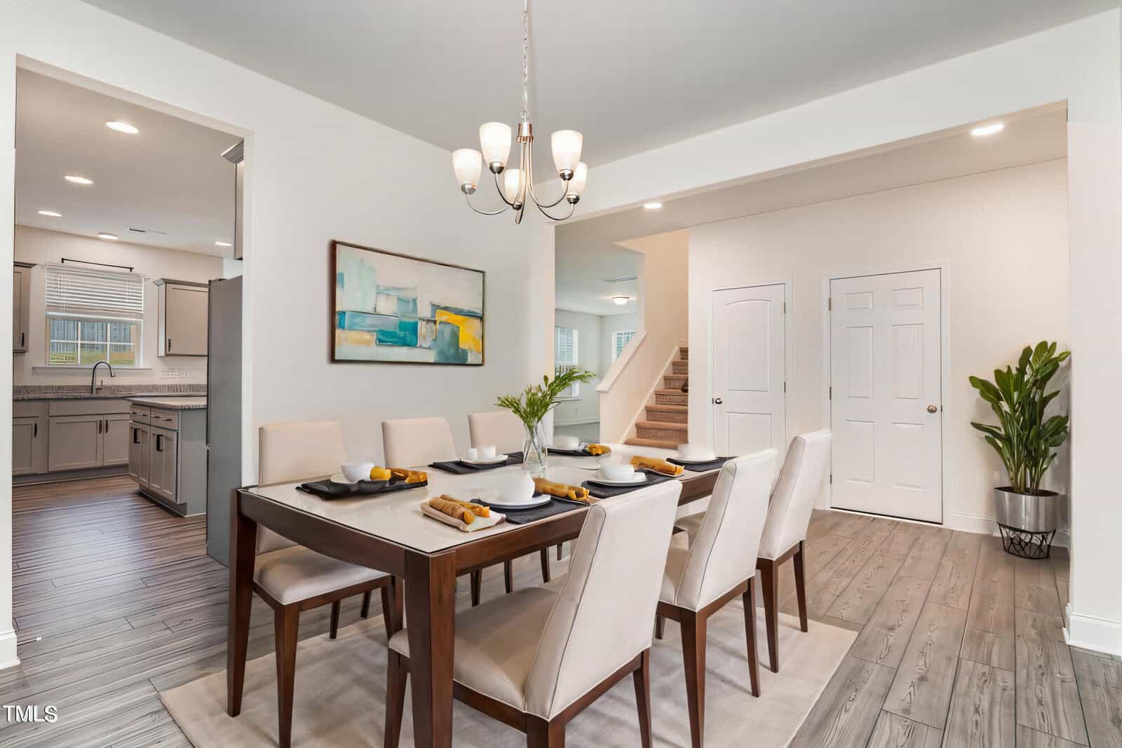 A modern dining room with a wooden table set for six, white upholstered chairs, a chandelier, abstract wall art, and a potted plant. The space opens to a kitchen and staircase with light-colored walls and flooring.