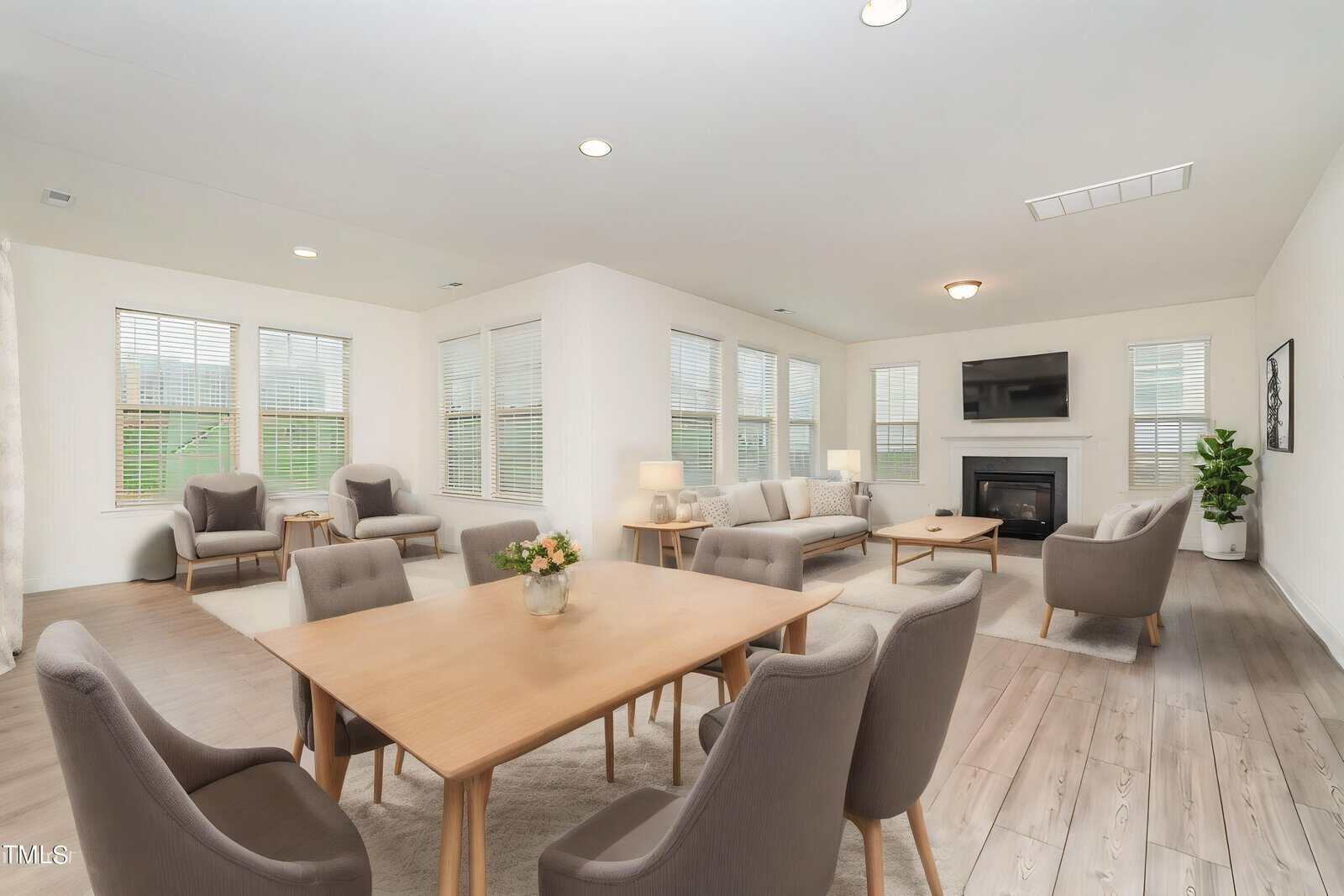 A spacious, modern living and dining area with light wood floors, neutral-colored furniture, large windows, a fireplace with a TV above it, and a potted plant in the corner.