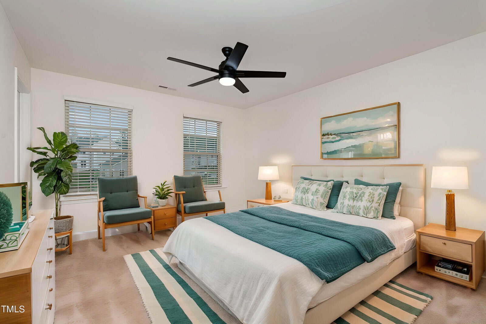 A modern bedroom with a large bed, teal and white bedding, two armchairs, nightstands with lamps, a ceiling fan, a striped rug, and a large framed artwork above the bed. Two windows let in natural light.