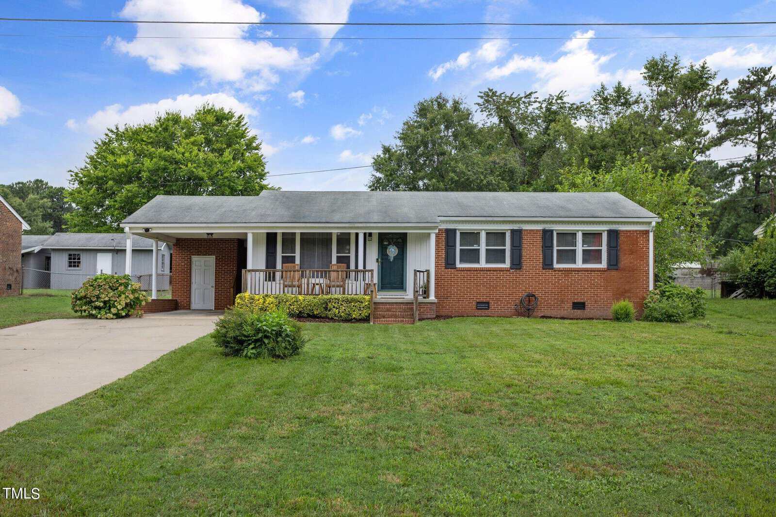A single-story brick house with a covered front porch featuring white railings and a dark green door, surrounded by a well-maintained green lawn. The driveway is to the left, leading to an attached carport. Tall trees are visible in the background under a partly cloudy sky.