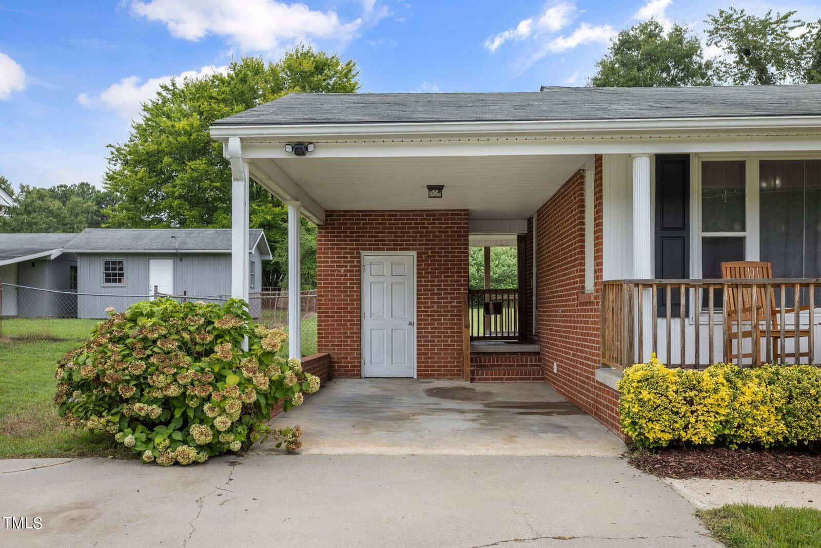 A brick house with a covered front porch and an attached carport. The porch has yellow bushes and a wooden rocking chair. The left side has a small storage room with a door. The background features a fenced yard with trees and a small shed.