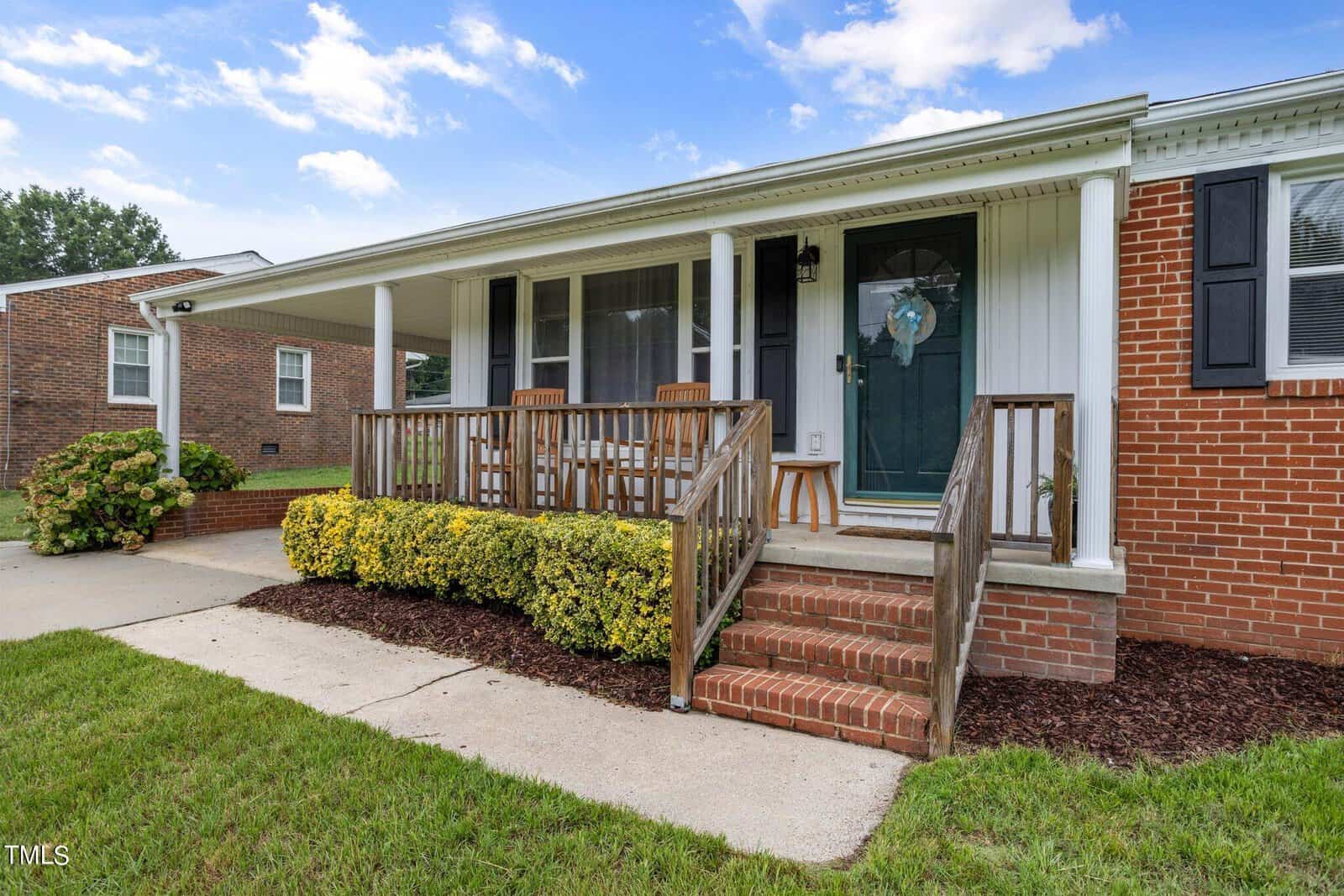 A single-story brick house with a covered front porch. The porch has a green door with a decorative wreath, wooden railings, two wooden chairs, and yellow shrubbery along the edge. A concrete pathway leads to the steps, and the lawn is neatly manicured.
