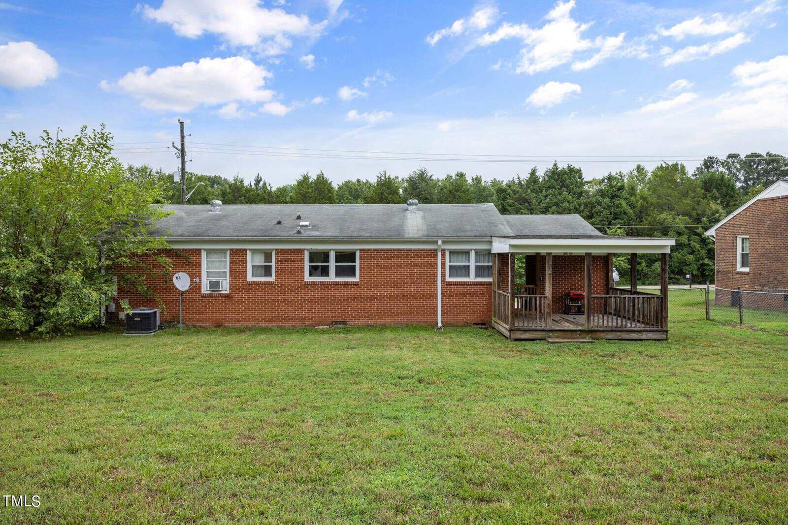 A brick house with a white roof sits on a grassy yard. It has a small covered porch with a red chair and multiple small windows. A satellite dish is attached to the side. A wooden fence and trees are visible in the background under a partly cloudy sky.