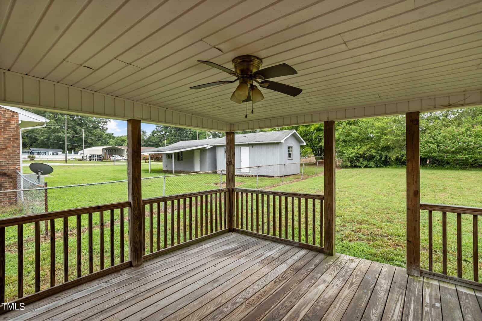Covered wooden deck with a ceiling fan, surrounded by railing. Overlooks a grassy yard with a chain-link fence. A small gray shed is visible in the background, along with trees and neighboring buildings.