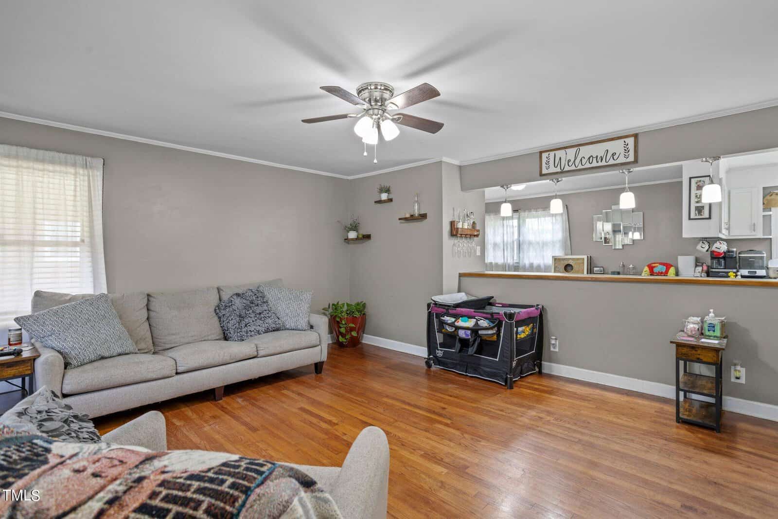 A living room with wooden floors, a gray sofa, and a ceiling fan with lights. A counter with pendant lights and various decorations is visible. A playpen is against the counter, and a plant is placed in a corner. A "Welcome" sign hangs above.
