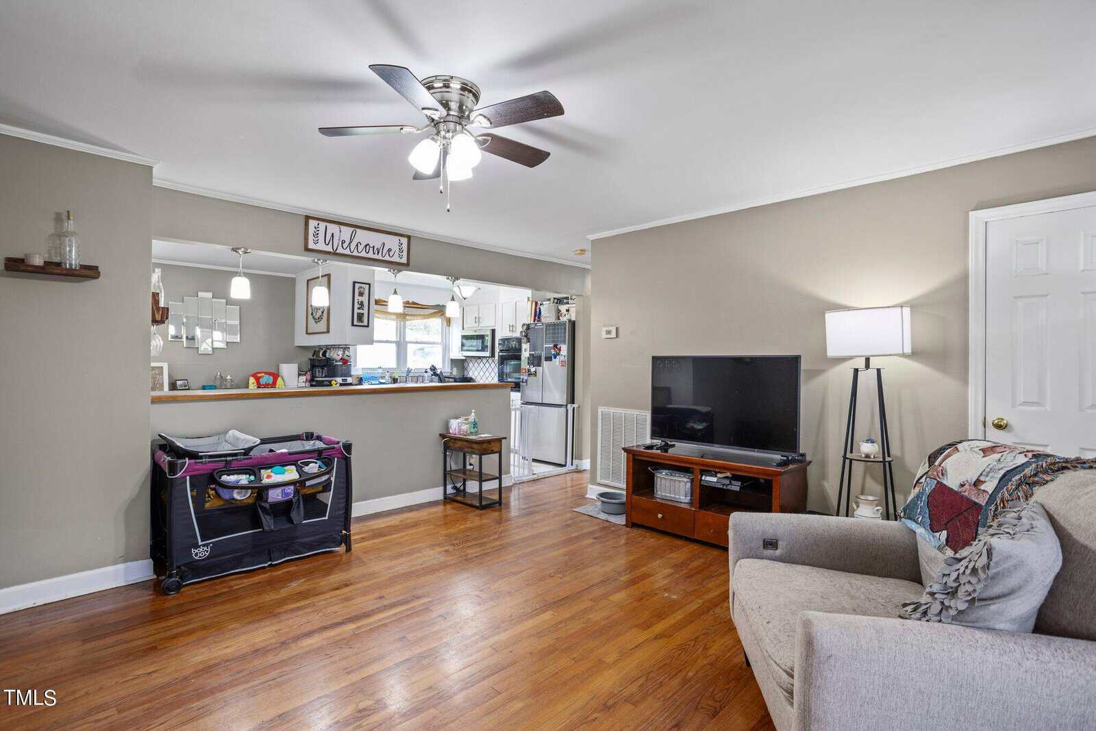 Living room with hardwood floor, beige walls, and ceiling fan. A TV and lamp are near a beige sofa with a patterned throw. A playpen is beside the kitchen counter in the background, which has pendant lights and a "Welcome" sign.
